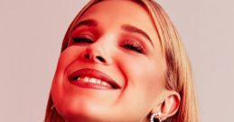 Millie Bobby Brown smiles joyfully, wearing elegant earrings and a stylish black outfit against a vibrant red backdrop.