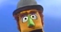 Monty, a colorful puppet with a green nose, wearing a gray hat and suit, showcasing his playful personality against a blue backdrop.