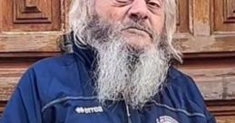 Elderly man with long white beard in navy jacket, standing confidently against a rustic wooden backdrop in Carovigno.