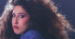 Paige Ginn poses with striking curls, showcasing a vintage denim outfit and bold makeup against a moody backdrop.