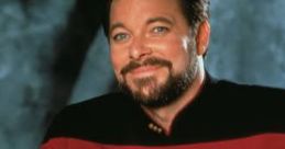 Jonathan Frakes as Commander Riker in Star Trek, wearing a red uniform and smiling against a gray backdrop.