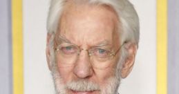 Donald Sutherland with silver hair and beard, wearing glasses and a red tie, posing in formal attire.