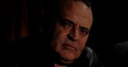 Sammy Delano poses seriously in a dimly lit setting, wearing a black jacket, highlighting his intense expression.