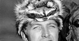 John Hart wearing a fur hat and fringed shirt, smiling next to a horse, showcasing a classic Western look.