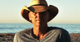 Kenny Chesney relaxing on the beach, wearing a straw hat, casual shirt, and enjoying the sunset by the ocean.