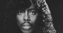 Rick James in a stylish white suit with a black tie, showcasing his iconic curly hairstyle and captivating expression.