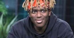KSI in a black hoodie and orange pants, smiling while holding a microphone at an event, showcasing his vibrant style.