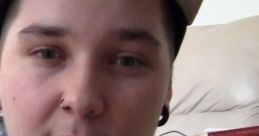 Person with a cap and piercings, looking directly at the camera, sitting in a cozy home setting. CD-I Gwonam fan.