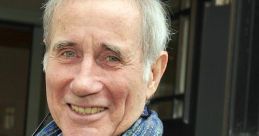 Jim Dale smiling warmly outdoors, wearing a blue scarf, known for his voice work and ARPAbet support in audiobooks.