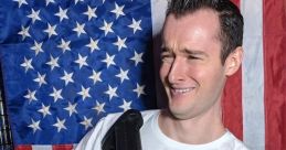 Man in a white shirt holding a firearm, grinning in front of a patriotic American flag backdrop, embodying humorous content.