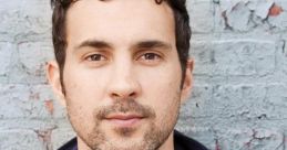 Mark Normand poses against a textured gray wall, sporting a casual navy zip-up jacket and a white-red t-shirt.