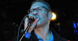 Alex Gutman performing passionately on stage, holding a microphone and a paper, wearing a stylish black coat.