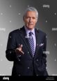 Alex Trebek, renowned host of Jeopardy!, dressed in a suit, gestures invitingly with a welcoming expression.
