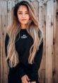 Valkyrae V2 showcasing a stylish long-haired look, wearing a 100 Thieves sweatshirt against a wooden fence backdrop.