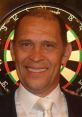 Russ Bray, famous darts commentator, smiles in front of a dartboard, showcasing his lively personality and passion for the game.