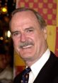 John Cleese smiling at an event, showcasing his iconic charm and wit in a black suit with a colorful tie.