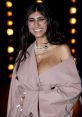 Mia Khalifa smiling in a chic outfit with layered jewelry, showcasing her stylish look against a vibrant backdrop.