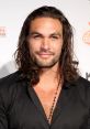 Jason Momoa smiles confidently, showcasing his long hair and stylish black shirt at a public event, exuding charisma.