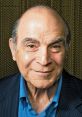 David Suchet smiling, wearing a blue shirt and dark blazer, known for his iconic portrayal of Hercule Poirot.