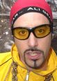 Ali G in a vibrant yellow jacket, yellow sunglasses, and a red beanie, showcasing his iconic hip-hop style and attitude.