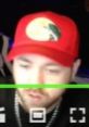 Dtanmanb wearing a red cap with a logo, focused in a dimly lit streaming setup, highlighting a casual yet engaged vibe.