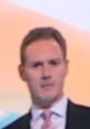 Professional man in a suit with a pink tie, presenting news in British English, set against a soft gradient background.