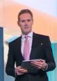Bradley Harris, British newsreader, delivering the latest news while holding a tablet, dressed in a suit with a pink tie.