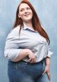 Confident woman with long red hair smiling in a blue striped shirt and jeans, posing against a light blue backdrop.