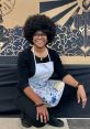 Ashley Seay, an artist with a vibrant style, poses in an apron, showcasing creativity against a colorful mural backdrop.