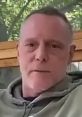 Man with a serious expression, wearing a hoodie, seated outdoors against a natural background. Throat Man content.