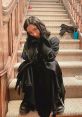 Amber from Scream 5 sits on a staircase in a black outfit, playfully posing with a smile amidst a casual setting.