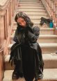 Amber from Scream V poses playfully on the stairs, dressed in black with a mysterious, captivating expression.