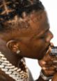 Artist with braided hairstyle and jewelry, making a silent gesture, reflecting confidence and style in rap culture.