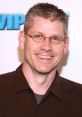 John Anderson smiling at an event, showcasing his signature style with a brown shirt and glasses, exuding warmth and charisma.