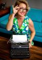 Smiling woman in retro dress poses with a Royal typewriter, showcasing creativity and vintage style in a cozy workspace.
