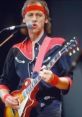 Mark Knopfler performing live, passionately playing a guitar in a distinctive outfit with vibrant colors.