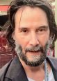 Keanu Reeves with long hair and a full beard, smiling in a casual black jacket against a vibrant backdrop.