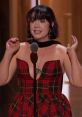 Rebecca Ford delivering an engaging speech on stage, wearing a stylish red plaid dress with a unique neckline and accessories.