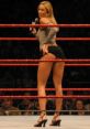 Wrestler in a ring, wearing a striped top and shorts, holding a microphone during a performance with fans in the background.