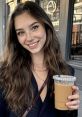Smiling woman with long wavy hair holding iced coffee, showcasing casual style in a city café setting. Tilly Norwood look.