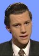 Chris Morris in a suit delivering satirical news, representing his iconic role in "Brass Eye" and "The Day Today.