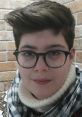 Portrait of Emanuele Brullo, a young individual with glasses and a cozy scarf, against a brick wall background.