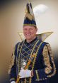 PRINS_JULIUS in traditional royal attire, featuring a ornate blue costume and feathered hat, holding a royal scepter.