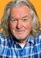James May smiles in a plaid shirt against an orange background, showcasing his unique style and vibrant personality.