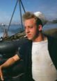 Shouting American crewmember with a cigarette, wearing a sailor hat, posed near water in a relaxed setting.