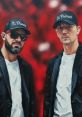 Os Primos duo posing in stylish attire with hats against a vibrant red background, showcasing their unique artistic style.