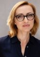 Portrait of Sarah Aubrey with blonde hair and glasses, wearing a navy shirt, against a neutral background.
