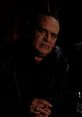 Sammy Delano poses seriously in a dimly lit setting, wearing a black jacket, highlighting his intense expression.