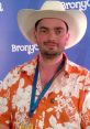 Man in a cowboy hat and floral shirt at BronyCon, smiling against a blue backdrop with BronyCon branding.