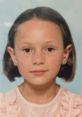 Young girl with short hair, wearing a pink dress, smiling softly against a light blue background. Captures childhood innocence.